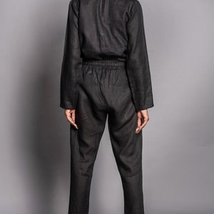 May include: A black long-sleeved jumpsuit featuring a collared neckline, a button-down front, and an elasticized waistband. The garment has tapered pant legs that end above the ankle, paired with beige ankle boots with a block heel.