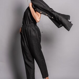 May include: A black sleeveless top and matching high-waisted trousers with an elastic waistband. A black jacket with light-colored buttons is draped over one shoulder. The outfit is paired with tan ankle boots featuring a side buckle and block heel.