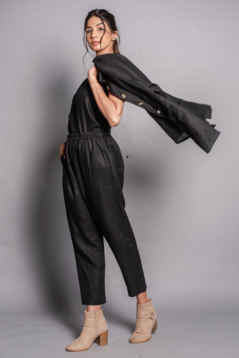 May include: A black sleeveless top and matching high-waisted trousers with an elastic waistband. A black jacket with light-colored buttons is draped over one shoulder. The outfit is paired with tan ankle boots featuring a side buckle and block heel.