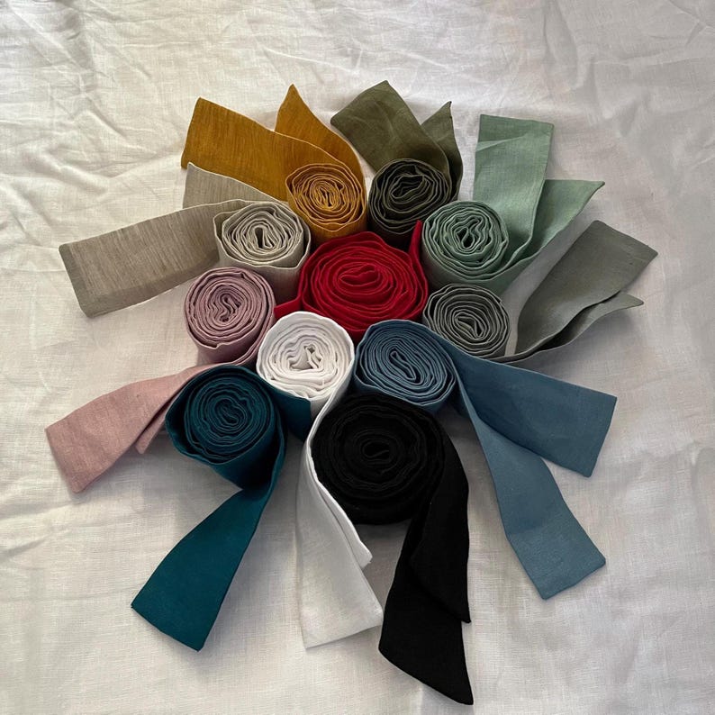 K&ouml;nnte beinhalten: Eine Sammlung von Stoffstreifen, die zu kreisf&ouml;rmigen Rollen gewickelt und in einem Kreis angeordnet sind. Die Farben umfassen Senfgelb, Olivgr&uuml;n, Salbei, Rot, Wei&szlig;, Schwarz, Altrosa, Petrol, Marineblau sowie verschiedene Grau- und Blaut&ouml;ne.