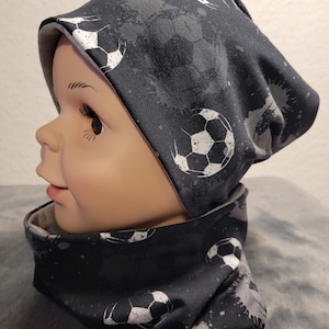 Könnte beinhalten: Ein schwarz-weiß gemustertes Mützen- und Halstuch-Set mit einem Fußball-Design. Die Mütze hat einen umgeschlagenen Rand und der Halstuch ist ein Schlauchmodell.