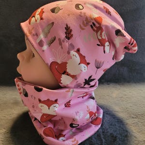 Könnte beinhalten: Rosa gestrickte Mütze und passender Halsschmuck mit Fuchs- und Blattmotiv.