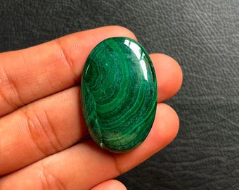May include: Oval-shaped malachite gemstone with a polished, glossy surface. The gemstone displays a rich, deep green color with swirling patterns. The stone is held in a hand, showcasing its natural beauty and unique veining.