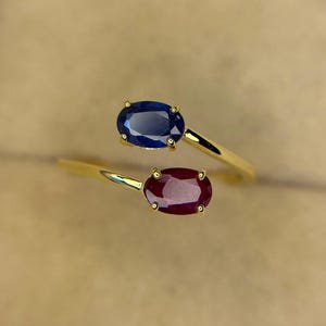 Puede incluir: Un anillo de oro tipo bypass con dos piedras preciosas de corte ovalado. Una piedra es un zafiro azul intenso y la otra es un rubí rojo oscuro. Ambas piedras están sujetas por engastes de cuatro garras en una banda de oro fina y pulida.