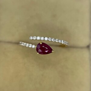 Op de afbeelding: Een open ring van geelgoud met een peervormige, dieprode robijn aan het ene uiteinde en een rij kleine, heldere, rondgeslepen witte diamanten langs de ringband, wat zorgt voor een verfijnd en elegant sieraad voor aan de vinger.