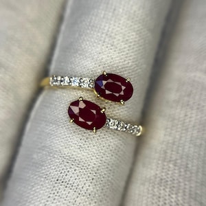 Puede incluir: Un anillo de estilo bypass con dos rubíes de color rojo intenso en talla oval engastados en oro amarillo. Cada extremo de la banda del anillo está acentuado con una hilera de pequeños diamantes blancos redondos, creando un brillo contrastante.