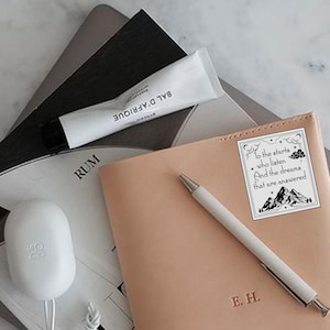 May include: A light brown leather journal with a white sticker on the front. The sticker has a black and white illustration of mountains and clouds with the text "To the stars who listen And the dreams that are answered". The journal also has the initials "E.H." embossed on the front.