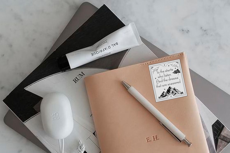 May include: A light brown leather journal with a white sticker on the front. The sticker has a black and white illustration of mountains and clouds with the text "To the stars who listen And the dreams that are answered". The journal also has the initials "E.H." embossed on the front.