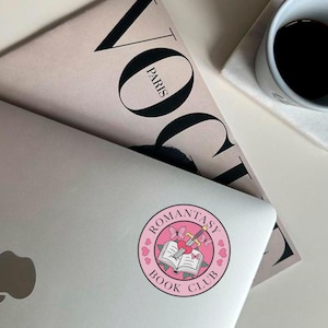 May include: A pink and white sticker with the text "Romantasy Book Club" in a circle. The sticker features a sword and a book with a heart on top.