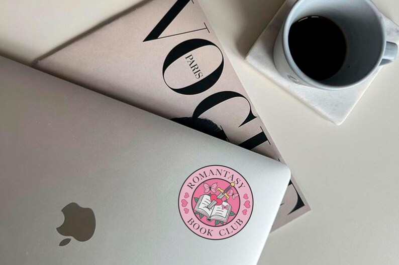 May include: A pink and white sticker with the text "Romantasy Book Club" in a circle. The sticker features a sword and a book with a heart on top.
