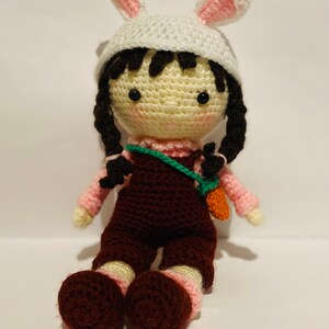 May include: A crocheted doll wearing a white bunny hat with pink ears, a pink shirt, brown overalls, and a green strap with a carrot attached. The doll has black hair and brown shoes.