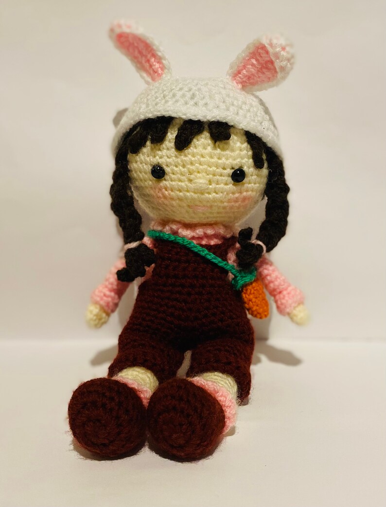 May include: A crocheted doll wearing a white bunny hat with pink ears, a pink shirt, brown overalls, and a green strap with a carrot attached. The doll has black hair and brown shoes.