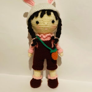 May include: A crocheted doll wearing a pink bunny hat, a pink shirt, brown overalls, and brown shoes. The doll is holding a green and orange bag.