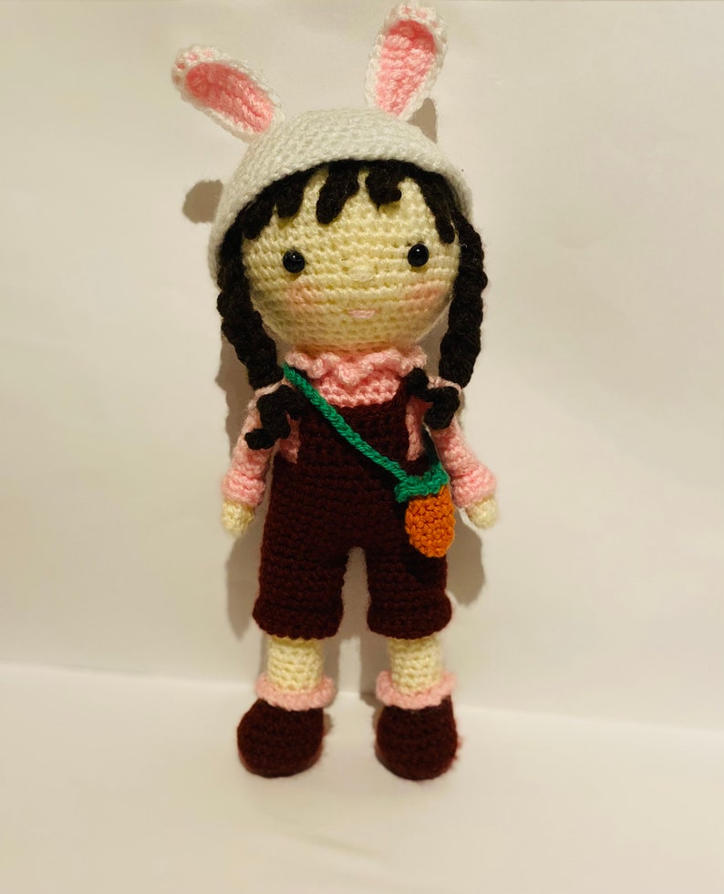 May include: A crocheted doll wearing a pink bunny hat, a pink shirt, brown overalls, and brown shoes. The doll is holding a green and orange bag.