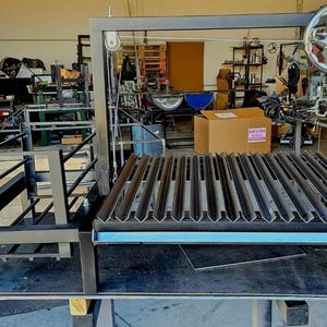 Può includere: Un grande barbecue in metallo costruito su misura con un telaio nero e una serie di griglie parallele. Il barbecue è dotato di un meccanismo a ruota ed è allestito in un ambiente di officina. Il design suggerisce che sia per cucinare all'aperto.