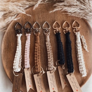 Könnte beinhalten: Sechs handgefertigte Schlüsselanhänger mit herzförmigen Metallverschlüssen und braunen Papieretiketten mit der Aufschrift "Handmade with Love". Die Schlüsselanhänger sind aus weißem, rosa, braunem, beigefarbenem, schwarzem und weißem Makramee-Schnur hergestellt. Ein Schlüsselanhänger hat eine weiße Makramee-Schnur mit dem Wort "MAMA" in Holzperlen.