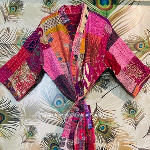 Könnte beinhalten: Eine rosafarbene und violette Patchwork-Kimono-Robe mit einem langen Bindegürtel. Die Robe ist aus einem weichen, leichten Stoff gefertigt und hat eine lockere Passform.