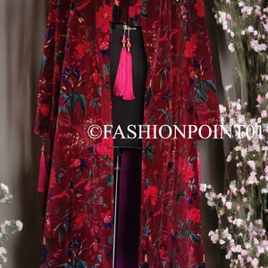 May include: A long, burgundy velvet kimono with a floral print. The kimono has a deep V-neck and long sleeves. It is open in the front and has a pink lining. The kimono is on a mannequin and has tassels on the sleeves.