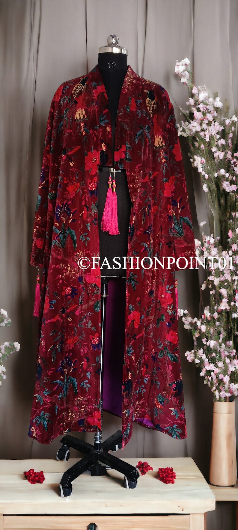 May include: A long, burgundy velvet kimono with a floral print. The kimono has a deep V-neck and long sleeves. It is open in the front and has a pink lining. The kimono is on a mannequin and has tassels on the sleeves.