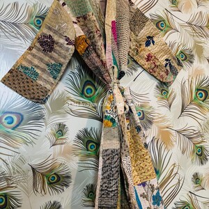 Puede incluir: Una bata acolchada de patchwork con una variedad de colores y patrones, incluyendo dise&ntilde;os florales, geom&eacute;tricos y paisley. La bata est&aacute; colgada de una percha sobre un fondo de plumas de pavo real.