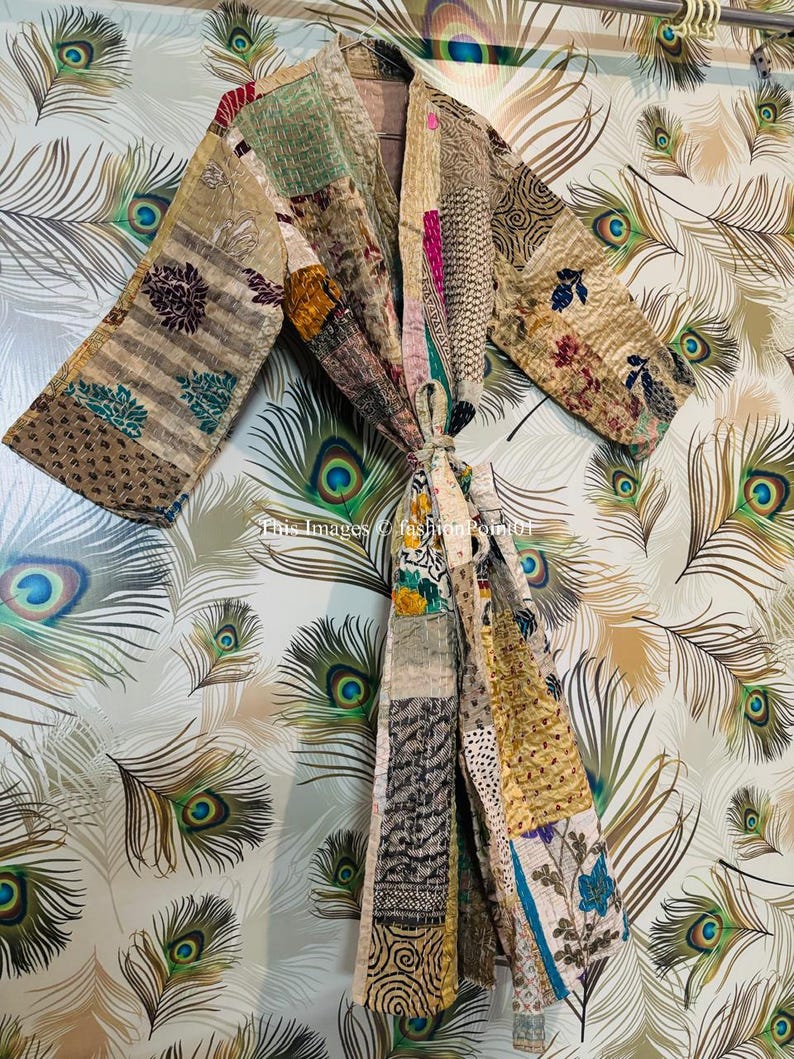 Puede incluir: Una bata acolchada de patchwork con una variedad de colores y patrones, incluyendo dise&ntilde;os florales, geom&eacute;tricos y paisley. La bata est&aacute; colgada de una percha sobre un fondo de plumas de pavo real.