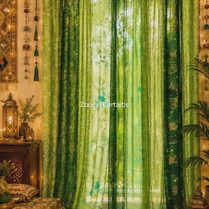 Könnte beinhalten: Transparente grüne Vorhänge mit verschiedenen botanischen und floralen Mustern in mehreren Grüntönen, aufgehängt mit Schlaufen und akzentuiert durch eine Schnur mit kleinen Quasten. Der Text Zooey Curtaibs ist mittig auf den Stoff gedruckt.