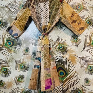 Puede incluir: Una bata de patchwork de estilo kimono con motivos en patchwork de colores marr&oacute;n, beige y verde. La bata tiene un cierre de nudo y mangas largas. La bata est&aacute; fotografiada sobre un fondo de plumas de pavo real.