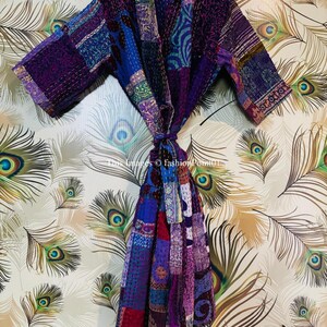 Op de afbeelding: Een patchwork kamerjas met diverse stoffen panelen in paars, blauw en magenta, voorzien van gedetailleerd kantha-stiksel. Het kledingstuk heeft lange mouwen, een sjaalkraag en een bijpassende stoffen ceintuur in de taille.