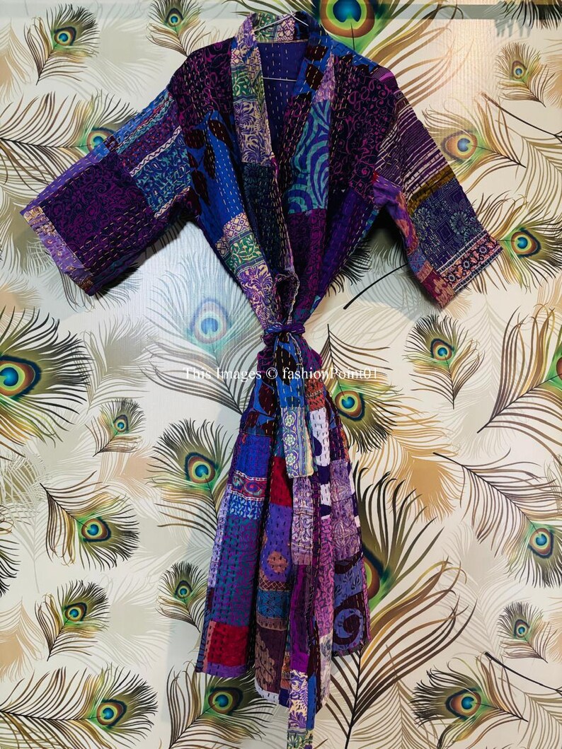 Op de afbeelding: Een patchwork kamerjas met diverse stoffen panelen in paars, blauw en magenta, voorzien van gedetailleerd kantha-stiksel. Het kledingstuk heeft lange mouwen, een sjaalkraag en een bijpassende stoffen ceintuur in de taille.