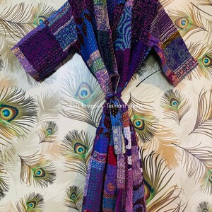 Op de afbeelding: Een kamerjas met lange mouwen en een patchworkontwerp in diverse tinten paars, blauw en roze. Het kledingstuk is opgebouwd uit meerdere stoffen panelen met zichtbare witte stikseldetails en een bijpassende ceintuur om te strikken.