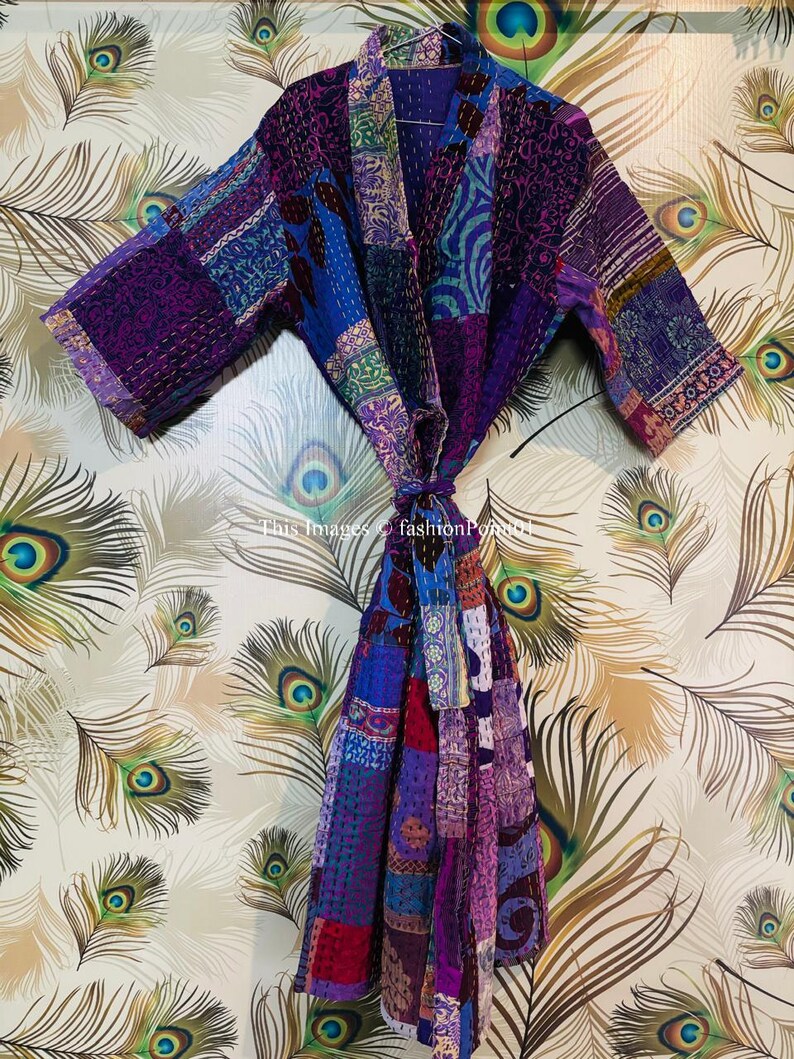 Op de afbeelding: Een kamerjas met lange mouwen en een patchworkontwerp in diverse tinten paars, blauw en roze. Het kledingstuk is opgebouwd uit meerdere stoffen panelen met zichtbare witte stikseldetails en een bijpassende ceintuur om te strikken.