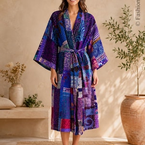 May include: A knee-length kimono-style robe featuring a patchwork design in various shades of blue, purple, and magenta. The garment has wide sleeves, a V-neckline, and a matching fabric tie belt at the waist, with visible white stitching throughout.