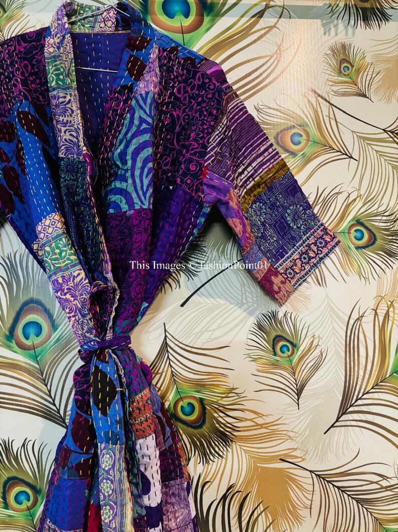 Op de afbeelding: Een patchwork kamerjas met een levendige mix van paarse, blauwe en magentakleurige stoffen panelen met ingewikkelde kantha-stiksels. Het kledingstuk heeft een sjaalkraag, driekwartmouwen en een bijpassende ceintuur. Tekst: This Images &copy;FashionPoint01.
