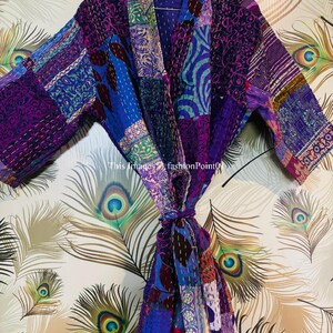 Op de afbeelding: Een kamerjas met lange mouwen en een patchworkontwerp in diverse tinten paars, blauw en roze. Het kledingstuk is opgebouwd uit meerdere stoffen panelen met zichtbare stikseldetails en is voorzien van een bijpassende ceintuur in de taille.