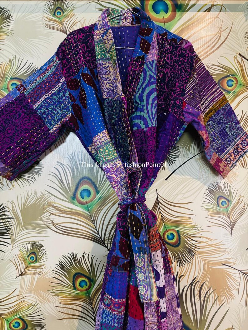 Op de afbeelding: Een kamerjas met lange mouwen en een patchworkontwerp in diverse tinten paars, blauw en roze. Het kledingstuk is opgebouwd uit meerdere stoffen panelen met zichtbare stikseldetails en is voorzien van een bijpassende ceintuur in de taille.