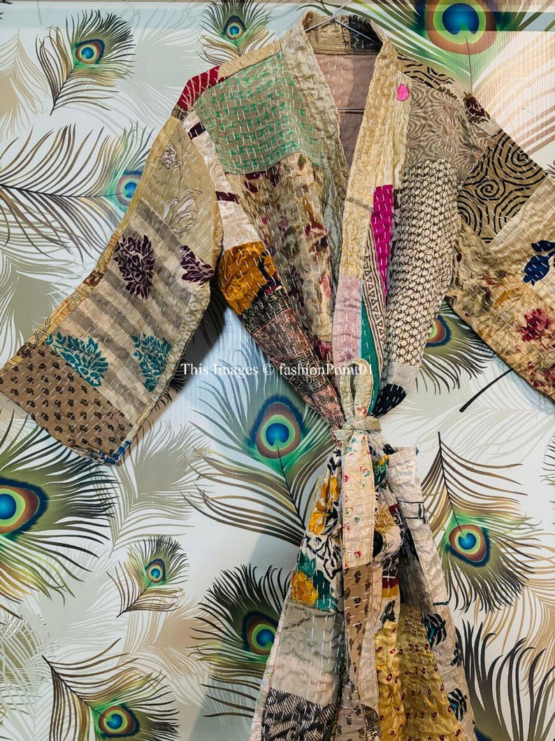 Puede incluir: Una bata de patchwork con una variedad de colores y patrones, incluyendo dise&ntilde;os florales, geom&eacute;tricos y paisley. La bata est&aacute; atada a la cintura con una cinta y tiene mangas largas. La bata est&aacute; fotografiada sobre un fondo de plumas de pavo real.