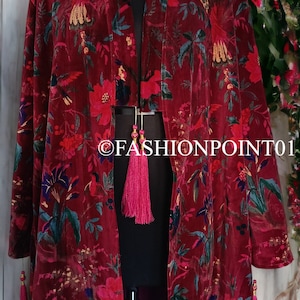 May include: A long, red velvet robe with a floral print featuring birds and flowers. The robe has a deep V-neckline and long sleeves with tassels at the cuffs and hem. The robe is lined with a purple fabric.