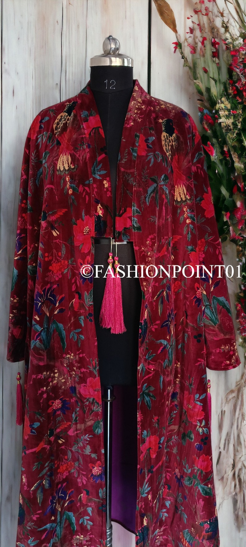 May include: A long, red velvet robe with a floral print featuring birds and flowers. The robe has a deep V-neckline and long sleeves with tassels at the cuffs and hem. The robe is lined with a purple fabric.