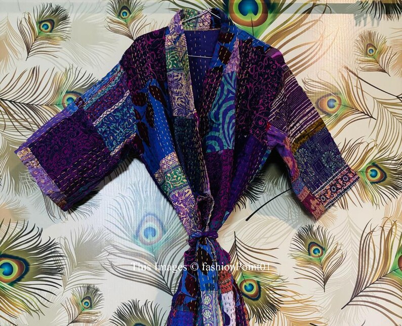 Op de afbeelding: Een patchwork kimono-badjas met diverse stoffen panelen in paarse, blauwe en donkere tinten met complexe patronen. Het kledingstuk heeft wijde mouwen, een sjaalkraag en een ceintuur, allemaal geaccentueerd met zichtbare witte kantha-stiksels.