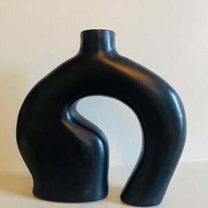 Könnte beinhalten: Eine schwarze Keramikvase mit einem einzigartigen, offenen Design. Die Vase hat einen schmalen Hals, der sich zu einem geschwungenen Körper mit einer großen Öffnung erweitert. Die Vase ist etwa 30 cm hoch und hat eine glatte, matte Oberfläche. Ein modernes Dekorationsstück.