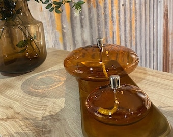 May include: Two amber glass oil lamps with a round, flat base and a silver wick holder. The lamps are sitting on a wooden surface.