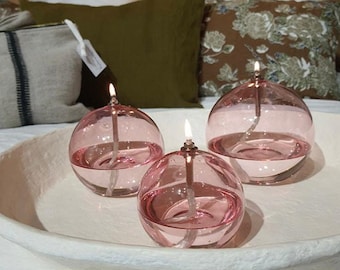 May include: Three pink glass oil lamps with wicks lit on a white tray. The lamps are round and have a clear glass base.