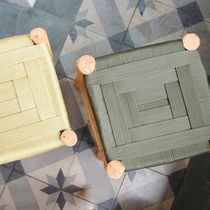 May include: Two wooden stools with woven rope seats. One stool has a light beige seat and the other has a dark green seat.