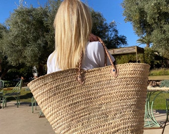 May include: A large, woven straw tote bag with braided handles and leather accents. The bag is carried over the shoulder. The natural beige colour of the bag contrasts with the blue sky and green foliage in the background.