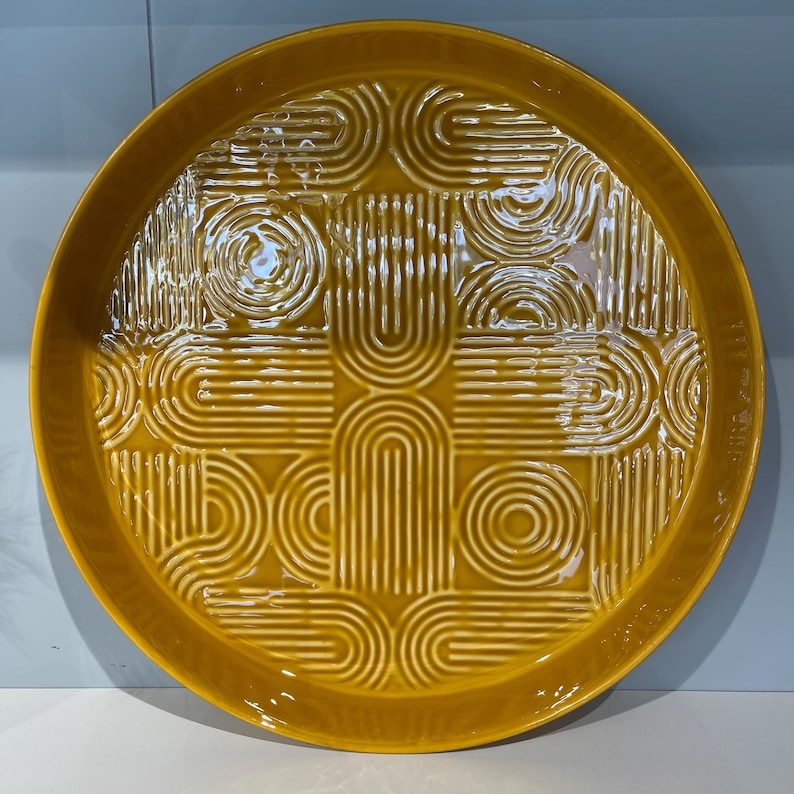 May include: A round, mustard-yellow ceramic serving platter with a raised geometric pattern. The design features repeating arcs, circles, and lines. The platter has a slightly raised edge and a glossy finish, suitable for serving food.