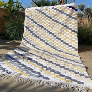 Peut inclure: Un tapis à carreaux blancs, jaunes et gris avec une bordure en franges. Le tapis est fait d'un matériau doux et moelleux et présente un motif géométrique.