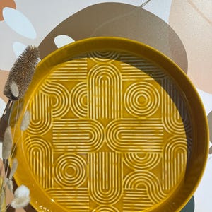 May include: A round, mustard-yellow ceramic tray with a raised geometric pattern of arches and circles. The tray has a slightly raised edge and a glossy finish. Decorative dried plants are visible on the left side of the image.