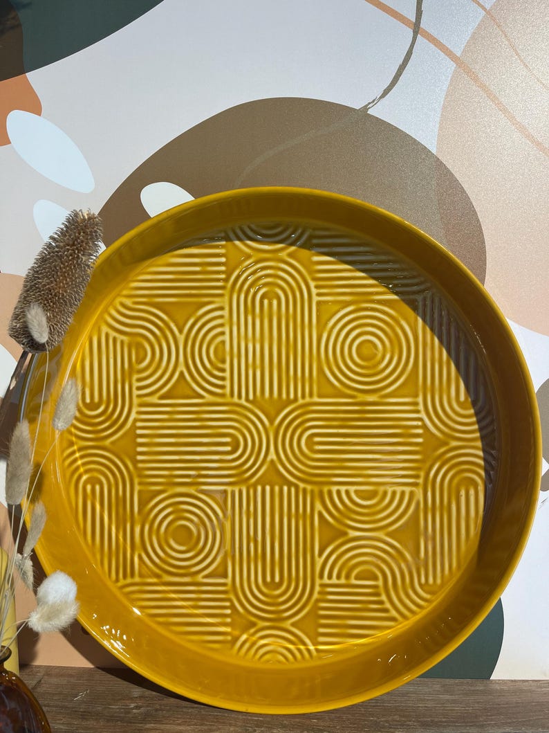 May include: A round, mustard-yellow ceramic tray with a raised geometric pattern of arches and circles. The tray has a slightly raised edge and a glossy finish. Decorative dried plants are visible on the left side of the image.