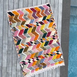 Peut inclure: Un tapis marocain coloré avec un motif géométrique de zigzags et de carrés dans des tons de rose, orange, jaune, vert, bleu et rouge. Le tapis a une bordure de franges blanches.