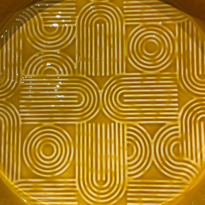 May include: Close-up of a yellow ceramic dish with a repeating geometric pattern. The design features curved lines and circles in a grid-like arrangement, creating a textured surface. The dish has a glossy finish.